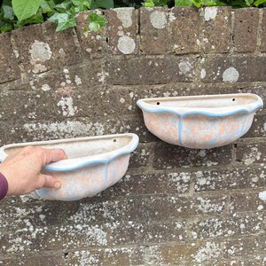 May include: Two wall-mounted ceramic planters with a scalloped edge and a pastel colour scheme. The planters have a peach base with blue accents and a blue trim. They are mounted on a weathered brick wall, suitable for flowers or small plants.