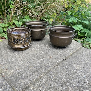 May include: Three brass plant pots, two with decorative designs, sitting on a grey concrete surface.