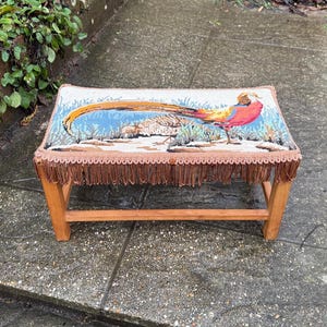 May include: A small wooden footstool with a tapestry top depicting a pheasant. The pheasant is brightly coloured with red, orange, and yellow plumage. The stool has a fringe around the bottom and measures approximately 40cm in length.