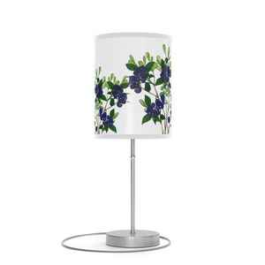 May include: A cylindrical table lamp with a white lampshade featuring a botanical design of blueberries and green leaves. The lamp has a silver metal base and stem, with a circular base and a visible power cord.