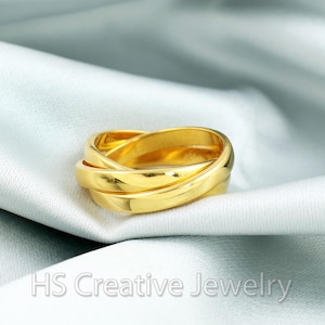 Può includere: Un anello dorato a incastro con tre fasce. L'anello è esposto su un tessuto azzurro chiaro. Il testo "HS Creative Jewelry" è visibile nella parte inferiore dell'immagine.