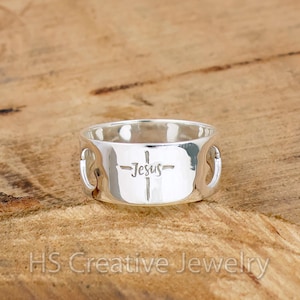 May include: A silver ring featuring the word "Jesus" engraved on a cross. The ring has a wide band with cut-out details. The ring is displayed on a wooden surface.