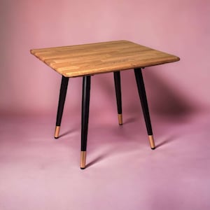 May include: A small square wooden table with black legs and gold accents. The table top is made of light wood and has a natural grain pattern.