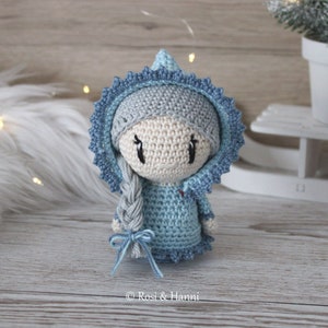Puede incluir: Una muñeca de crochet con un vestido con capucha azul y gris, cabello gris trenzado y un lazo azul. La muñeca tiene una cara blanca con ojos negros y un corazón rojo en el pecho.