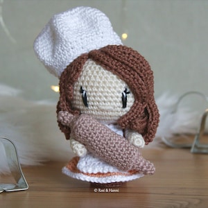 Puede incluir: Una muñeca de crochet con un gorro de chef blanco y sosteniendo un rodillo de amasar marrón. La muñeca tiene el pelo marrón y un vestido blanco con ribetes naranjas.