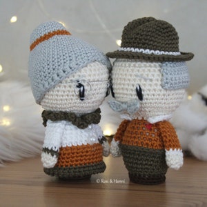 Puede incluir: Dos muñecas de crochet, un hombre y una mujer, de pie uno al lado del otro. La mujer lleva un sombrero gris, un suéter blanco y una falda marrón. El hombre lleva un sombrero marrón, un suéter marrón y pantalones verdes.  © Rossi & Hanni
