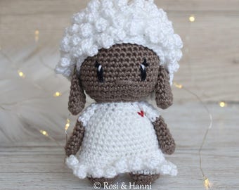May include: A handmade crochet lamb doll with a white, fluffy head and a white dress. The lamb has a brown face, ears, and limbs. A small red heart is stitched on the dress. The doll is on a light wood surface. The text "© Rosi & Hanni" is at the bottom.