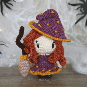 May include: A crocheted witch doll with a purple hat, orange hair, and a brown broom. The doll is wearing a purple dress with yellow accents and orange polka dots.