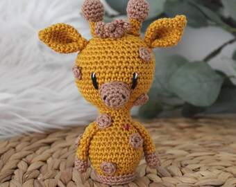 May include: A crocheted giraffe toy with brown spots and a red heart on its chest. The giraffe is standing on a woven surface.