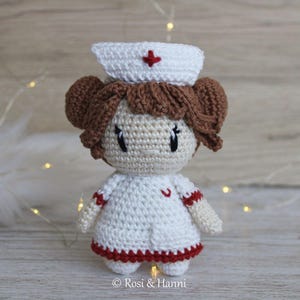 May include: A crocheted doll wearing a white nurse's uniform with red trim and a red cross on her hat. The doll has brown hair and a sweet expression.