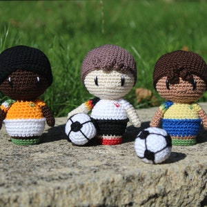 Puede incluir: Tres figuras de jugadores de fútbol de ganchillo, cada una con una camiseta diferente. Los jugadores sostienen balones de fútbol y están de pie sobre una superficie de piedra.