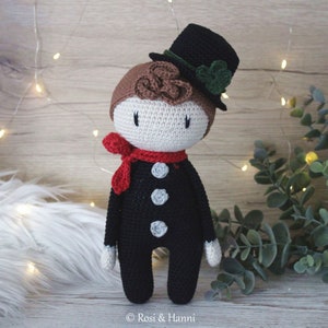 May include: A crocheted doll wearing a black suit, a red scarf, and a black top hat with green leaves. The doll has brown hair and a white face with black eyes.