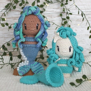May include: Two crocheted mermaid dolls with blue and green hair. One doll has brown skin and the other has white skin. Both dolls are wearing blue and green crocheted mermaid tails.