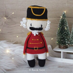 May include: A crocheted nutcracker doll with a red and gold uniform, white beard, and black hat. The doll is standing on a white surface with a small Christmas tree in the background.