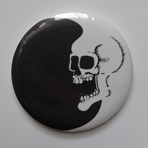 May include: A round, black and white button featuring a skull design. The skull is on the right side of the button, with the left side being black. The skull has an open mouth and is detailed with black lines.