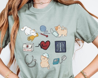 May include: A light green t-shirt with a graphic design featuring a variety of cat-themed medical illustrations, including a stethoscope, a first aid kit, a heart, an x-ray, a blood pressure cuff, a cat wearing a nurse's hat, and a cat with a bandage on its paw.