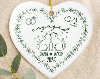 May include: A white heart-shaped ceramic ornament featuring a dark green floral border. The center displays the text engaged, two kissing cats, two rings, a bird with a heart, and the names Sharon and Jackson with the year 2026 below.