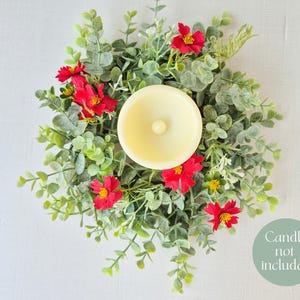 May include: A decorative candle ring featuring a cream-colored candle holder surrounded by green foliage and vibrant red flowers. The design includes small yellow accents and is set against a white background. The text "Candle not included" is visible.