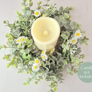 May include: A cream-colored candle sits in the center of a decorative wreath. The wreath is made of green leaves and small white daisies. The text "Candle not included" is on a green circle.