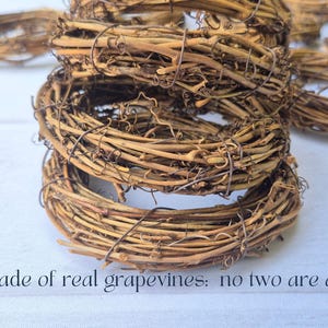May include: Several grapevine wreaths stacked on a white surface. The wreaths are made of natural brown vines, with a rustic and organic appearance. The text "Made of real grapevines: no two are alike" is visible.