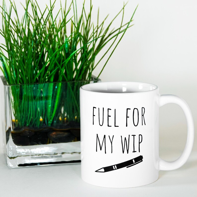 May include: White ceramic mug with the text "FUEL FOR MY WIP" in black, along with a black pen illustration. A clear glass square vase with green grass is in the background.