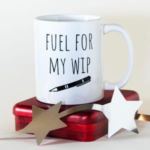 May include: White ceramic mug with the text "FUEL FOR MY WIP" in black, along with a black pen illustration. The mug sits atop a red rectangular tin, with a silver star-shaped decoration.