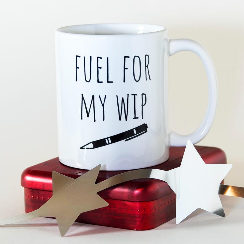May include: White ceramic mug with the text "FUEL FOR MY WIP" in black, along with a black pen illustration. The mug sits atop a red rectangular tin, with a silver star-shaped decoration.