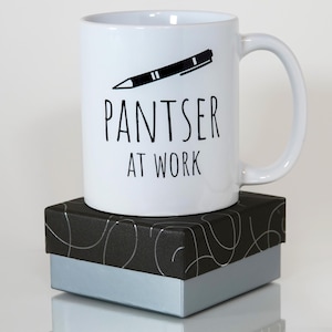 May include: White ceramic mug with a black pen illustration and the text "PANTSER AT WORK." The mug sits on a black and silver patterned gift box with a light blue base. The mug has a curved handle.