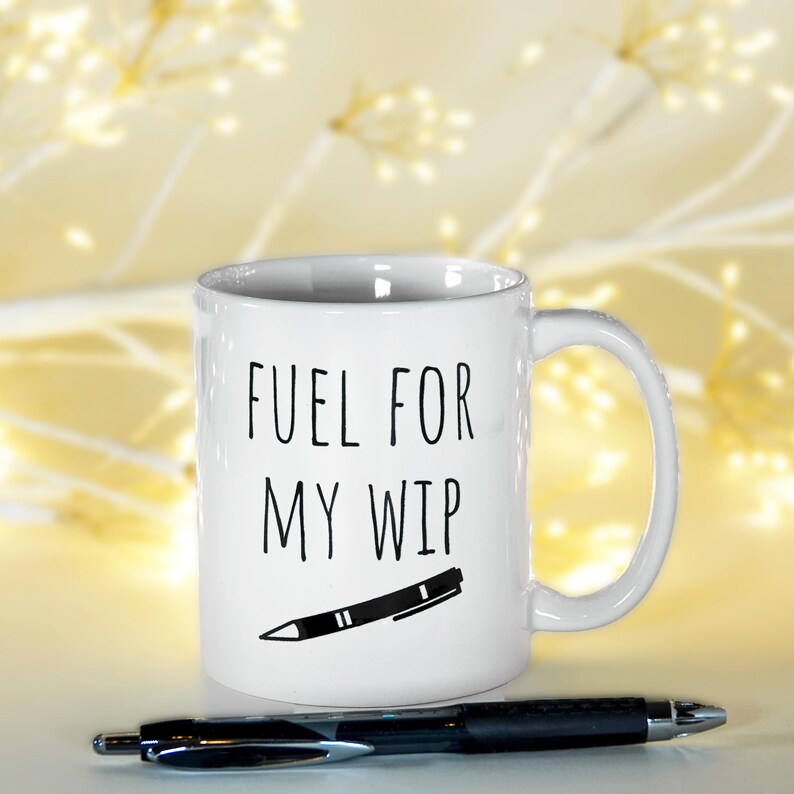 May include: White ceramic mug with the text "FUEL FOR MY WIP" in black, along with a black and white pen illustration. A black pen rests in front of the mug. The background is a soft, blurred yellow.