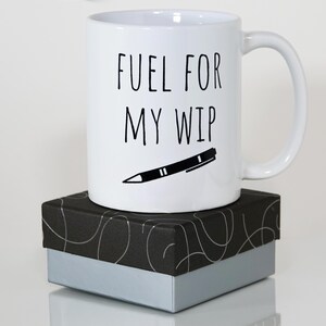May include: White ceramic mug with the text "FUEL FOR MY WIP" and a black pen illustration. The mug sits on a black and silver gift box. The mug has a curved handle.
