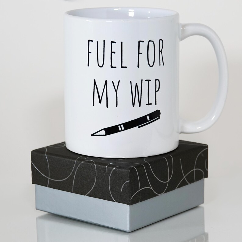 May include: White ceramic mug with the text "FUEL FOR MY WIP" and a black pen illustration. The mug sits on a black and silver gift box. The mug has a curved handle.