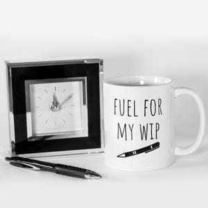 May include: A white coffee mug with the text "FUEL FOR MY WIP" and a pen illustration. A black pen and a small square clock with a black frame are also in the image.