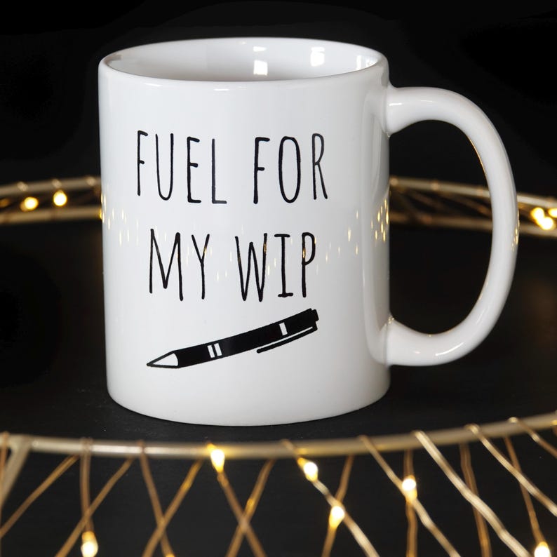 May include: White ceramic mug with the text "FUEL FOR MY WIP" in black, along with a black and white pen illustration. The mug has a curved handle and is set against a dark background.