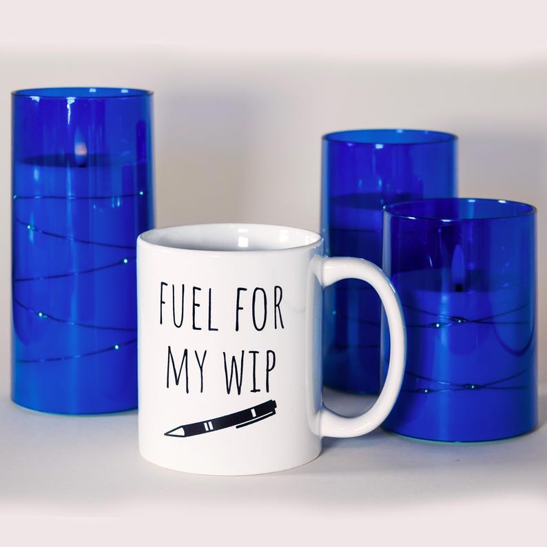 May include: A white ceramic mug with the text "FUEL FOR MY WIP" and a pen illustration. The mug is set in front of three blue glass candle holders with string lights.