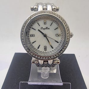 Puede incluir: Un reloj plateado con esfera blanca y la palabra "Papillon" impresa. El reloj tiene un bisel incrustado con pequeñas piedras transparentes y una correa plateada. El reloj se exhibe sobre un soporte transparente sobre un fondo blanco.