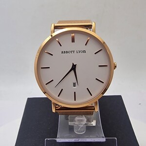 May include: Gold-coloured wristwatch with a white face and black hands. The watch features the brand name "abbott LYON" and a date display. The watch has a mesh strap and is displayed on a black stand.