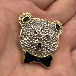Puede incluir: Broche vintage de oso de peluche con marco dorado. La cabeza del oso está cubierta de pequeños y brillantes pedrería. Tiene detalles de esmalte negro en las orejas y una pajarita negra. Los ojos del oso son rojos.