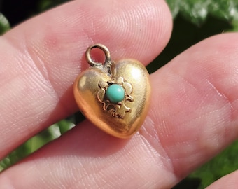 May include: A small, gold-colored heart-shaped pendant with a turquoise-colored stone in the center. The pendant has a small loop at the top for a chain. The heart is smooth and shiny, with a floral design around the stone.