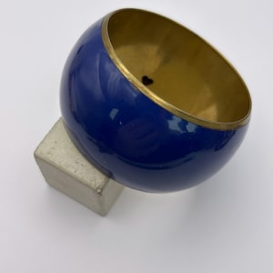 Puede incluir: Un brazalete azul marino con interior y ribete dorados. El brazalete tiene forma redondeada y descansa sobre un pequeño bloque rectangular gris. El brazalete es un accesorio de moda.