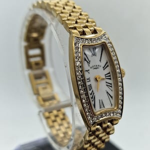 Può includere: Orologio da polso Rotary color oro con quadrante rettangolare e bracciale a maglie. Il quadrante è bianco con numeri romani neri e il marchio "Rotary". La lunetta è incastonata con piccoli cristalli trasparenti.