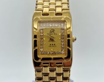May include: Gold-toned rectangular wristwatch with a matching link bracelet. The watch face is square, with a gold background, small diamonds, and the text "ROYMAD WUL" and "10M GOLD". The watch is displayed on a clear stand.