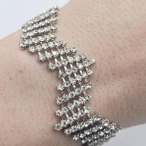 Op de afbeelding: Een zilverkleurige armband met een zigzagontwerp, versierd met talrijke heldere, sprankelende strass-steentjes. De armband wordt getoond op een pols, waarbij het ingewikkelde patroon en de reflecterende eigenschappen worden benadrukt. Geschikt voor formele gelegenheden.