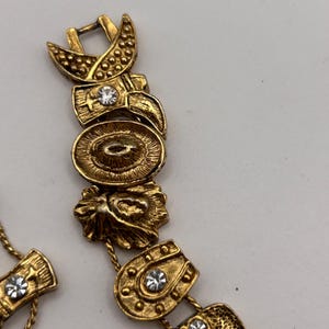 May include: Gold-toned vintage bracelet featuring various decorative elements. The bracelet includes crescent shapes, oval medallions, and floral designs, all adorned with small, clear rhinestones. The clasp is a simple hook and eye closure.