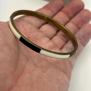 Puede incluir: Un brazalete dorado con segmentos de esmalte negro y blanco alternados. El brazalete tiene un diseño simple y circular, que muestra una combinación de colores clásica. Un accesorio elegante.