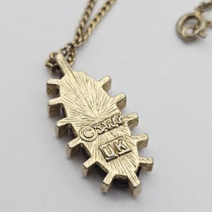 May include: A gold-toned necklace with a pendant featuring a unique, textured design. The pendant has a series of rectangular shapes with a sunburst pattern. The back of the pendant is stamped with the text "SARAH" and "UK".