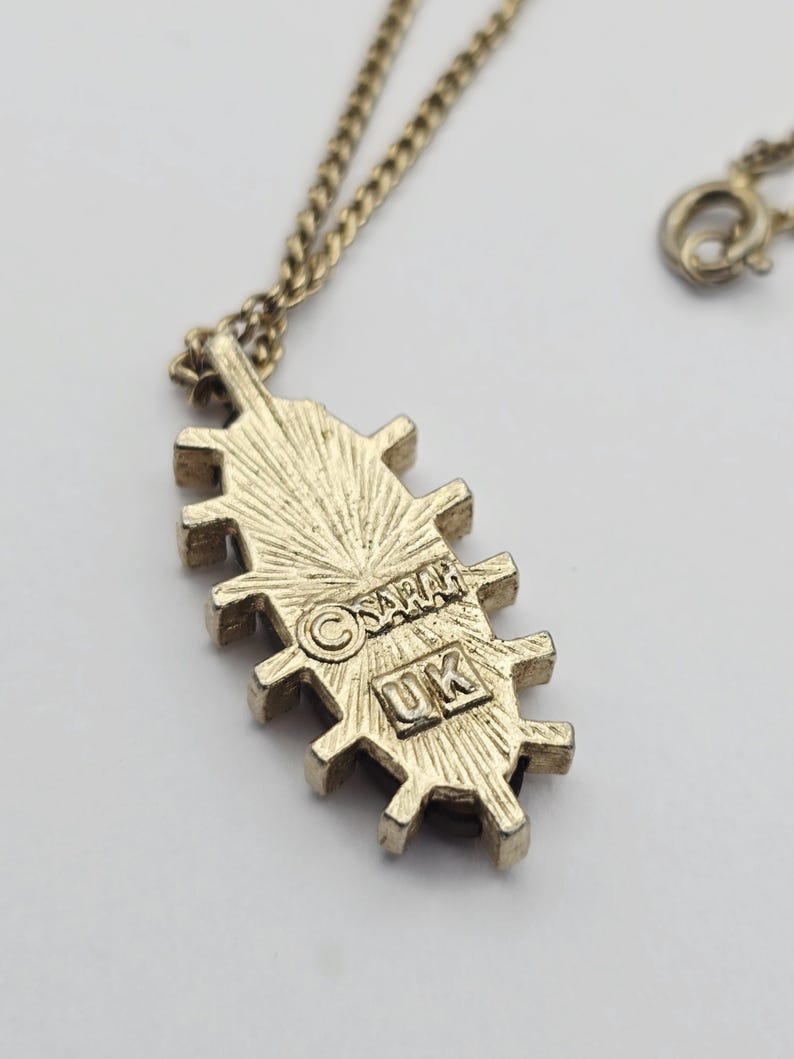 May include: A gold-toned necklace with a pendant featuring a unique, textured design. The pendant has a series of rectangular shapes with a sunburst pattern. The back of the pendant is stamped with the text "SARAH" and "UK".