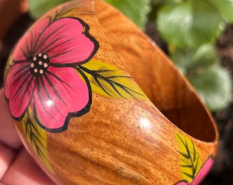 May include: Wooden bangle bracelet with a hand-painted floral design. The bracelet features a pink flower with black details and green leaves against a natural wood grain background. The bangle is approximately 3 inches in diameter.