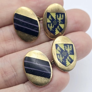 Puede incluir: Un par de gemelos dorados. Uno presenta un diseño a rayas negras y moradas, el otro un escudo azul y amarillo con figuras de pájaros. Los gemelos tienen forma ovalada.