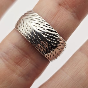 Può includere: Un anello d'argento con un motivo strutturato e inciso. L'anello è largo e presenta un disegno ripetuto di linee angolate. L'anello è mostrato su un dito su sfondo bianco.