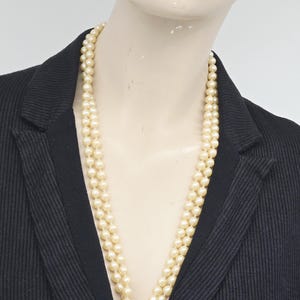 Può includere: Una collana di perle a due fili di colore crema. La collana è drappeggiata su un manichino che indossa un blazer nero. Le perle sono di dimensioni uniformi e creano un look classico ed elegante. Il blazer ha una sottile trama a coste.